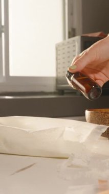 Dikey video. Kadın elinin, mum yapımında kullanılan aletlerin yanında, güneş ışığına karşı küçük bir kehribar yağı şişesi tutarken yakın plan. Aromaterapi kavramı
