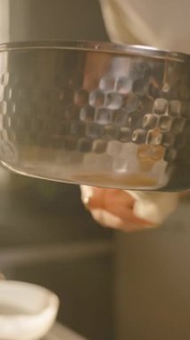 Dikey video. Kadın mutfaktaki mum kaplarının yanındaki masada eritilmiş balmumuyla saksıyı tutuyor. El Yapımı Mum Projesini Kabul Et