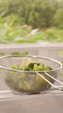 Dikey video. Colander Buharlı Brokoli ile Dolu Açık Pencere Ledge with Lush Garden in Background, Geniş Açı, Doğal Gün Işığı, Sağlıklı Aşçılık Konsepti