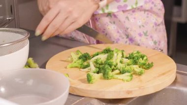 Çiçekli Önlüklü Kadın Mutfak Tezgahı, Parlak Aydınlatma, Yemek Hazırlama Konsepti Yuvarlak Tahta Üzerinde Taze Brokoli Kesiyor