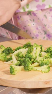 Dikey video. Çiçekli Önlüklü Kadın Mutfak Tezgahı, Parlak Aydınlatma, Yemek Hazırlama Konsepti Yuvarlak Tahta Üzerinde Taze Brokoli Kesiyor