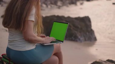 Kaya Üzerinde Oturan Kadın Deniz Kıyısı, yan görünüş, Uzaktan Çalışma Konsepti yakınlarındaki Yeşil Ekran ile Laptop kullanıyor