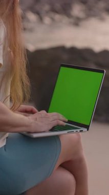 Dikey video. Kaya Üzerinde Oturan Kadın Deniz Kıyısı, yan görünüş, Uzaktan Çalışma Konsepti yakınlarındaki Yeşil Ekran ile Laptop kullanıyor