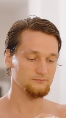 Dikey video. Banyodaki aynada alnına yüz kremi süren adam, yakın açı, erkek cildi kavramı.