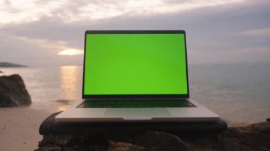 Yeşil ekranlı dizüstü bilgisayarın ön görüntüsü Gün batımına karşı kaya üzerinde duruyor.