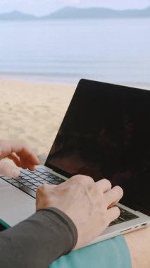 Dikey video. Sakin Okyanus Arkaplanı Uzaktan Kumandayla Tropikal Kumsal Sahili 'nde otururken Dizüstü bilgisayarda yazan Erkek Elleri' nin Kapanışı