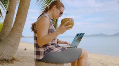 Tropikal Plajda oturan ve dizüstü bilgisayarla hindistan cevizi içen kadın Okyanus Ufku Egzotik Yaşam Tarzı
