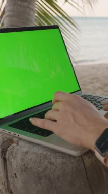 Dikey video. Eller dizüstü bilgisayarda, tahta gövde üzerinde yeşil ekranla, hindistan cevizi içeceğinin yanında, Ocean in Ocean in Close-Up Shot, Uzak Görev Konsepti ile yazılıyor.