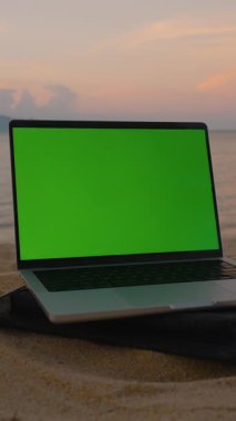 Dikey video. Kumsalda hindistan cevizi içeceğinin yanında yeşil ekranlı dizüstü bilgisayar Okyanus ve Günbatımı Arkaplanı geniş çekim, Dijital Göçebe Konsepti