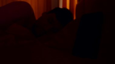 Yatakta yatan adam akıllı telefon alarmına doğru bakıyor. Sabah 6 'da penceresinden portakal ışığı süzülüyor.