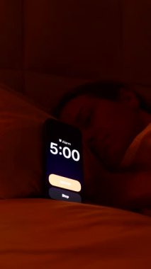 Dikey video. Sabahın 5 'inde yatakta uzanan, Smartphone Alarmı' nı kapatan, Parlak Pencereden dışarı bakan ve Sabah Tembelliği 'ni tekrar uykuya daldıran kadın.