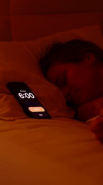 Dikey video. Sabah 6 'da beyaz çarşafların altında uzanmış, parlak pencereden dışarı bakan ve tekrar uykuya dalan kadın alarmını kapatıyor.