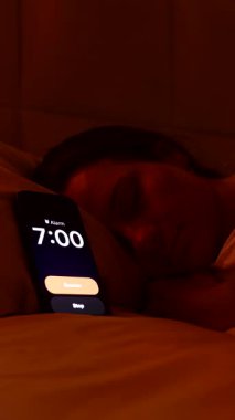 Dikey video. Beyaz çarşafın altında yatan kadın sabah 7 'de alarmı kapatıyor. Parlak pencereden dışarı bakıyor ve yine geç saatte uyuyakalıyor.