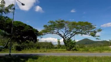 View from Moving Car of Small Rural House with Blue Roof among Green Trees and Mountains under Bright Blue Sky Countryside Journey