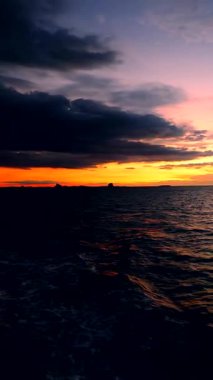 Vertical video. Vertical View of Fiery Orange Sunset Over Dark Ocean Waves with Heavy Clouds and Distant Silhouettes Tranquil Evening Voyage