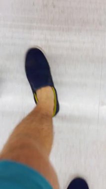 Vertical video. First-Person View of a Person Walking on a Light Floor Between the Wheels of a Shopping Cart in a Supermarket, Casual Outfit, Daily Routine Concept