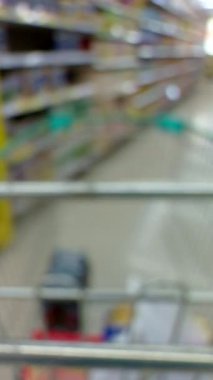 Vertical video. First-Person View Through an Empty Shopping Cart Moving Down a Bright Supermarket Aisle, Colorful Shelves Out of Focus, Consumerism Concept