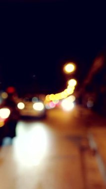 Vertical video. Blurred View of Car Driving on City Street at Night with Headlights Glowing and Streetlights Creating Bokeh Circles, Front Angle, Shallow Depth of Field, Night Traffic Concept