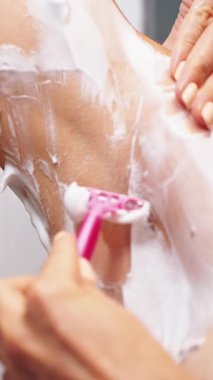 Vertical video. Woman Shaving Underarm Using Razor and Thick Shaving Cream in Bathroom, Arm Raised, Close-Up Shot, Personal Grooming Concept