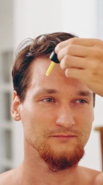 Dikey video. Sarı Retinol Serumunu Alnına Düşüren, Cilt Rutini ve Yaşlanma Karşıtı Kavram ile Uygularken Aynaya Bakan Adam