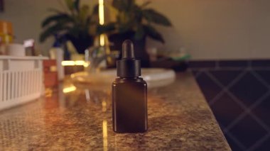 Hand Holding Small Dark Glass Bottle with Dropper Dispensing Yellow Liquid on Bathroom Countertop in Warm Evening Light, Skincare and Self Care Concept