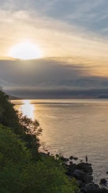 Vertical video. Green Cliff by Calm Sea Under Bright Sunlight and Blue Sky with Soft Cloud Layers at Sunset, Sunset Timelapse Concept