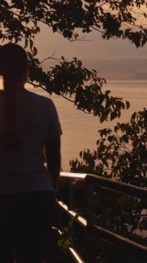 Dikey video. Ön planda Siluetli Yeşilliklerle Sahne Yolu 'nda Dururken Kadın Okyanus Günbatımına Bakar Sıcak Akşam Işığından Yakalandı