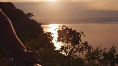 Güneş batımında okyanusa bakan metal korkuluklara yakın mesafede Tropikal yeşillik ve dağ siluetiyle çevrili ılık altın saat aydınlığında