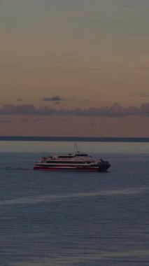 Vertical video. Ferry Boat Moves Gently Across Open Sea at Sunset Toward Horizon Surrounded by Faint Clouds and Calm Waters in Wide Angle View