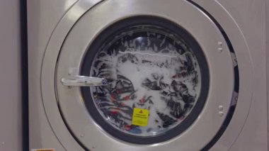 Close View of Laundry Machine Filled with Bubbly Water and Colorful Clothes Spinning during Wash Cycle, Captured from Front Angle, Cleaning Process Concept