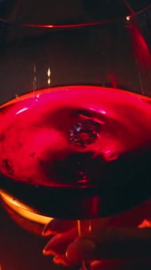 Vertical video. Hand Holding Glass of Red Wine Beside Candle with Deep Red Light Reflection on Surface, Cozy Night Relaxation Concept