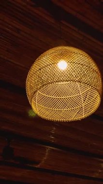 Dikey video. Akşam Ortamı Ahşap Tavanın Altında Asılı Örgü Bambu Lambanın Ilık Işığı, Tropikal Tasarım Konsepti ve Samimi Atmosfer