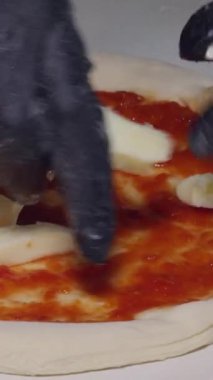 Dikey video. Ellerinde siyah eldivenli Mozzarella peyniri şeritleri ile pizza hamuruna domates sosu, ev yapımı yemek pişirme ve İtalyan mutfağı hazırlığı.