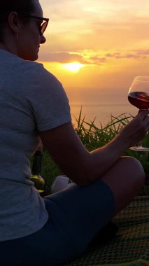 Dikey video. Çimenlerde oturan kadın elinde şarap kadehiyle okyanus günbatımını seyrediyor. Yan manzara, huzur dolu yalnızlık kavramı.