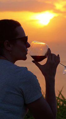 Dikey video. Mat 'in üzerinde otururken Şarap İçen Kadın Okyanus, Geniş Açı, Huzur Yalnızlığı Konsepti Üzerinde Gün Batımını İzliyor