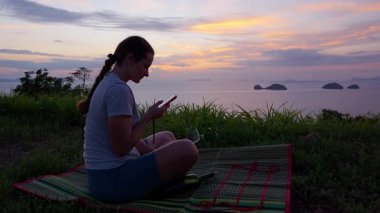 Mat 'te Oturan Kadın Okyanus, Geniş Açı, Rahat Yaşam Tarzı Konsepti Üzerinde Günbatımını izlerken Akıllı Telefon Kullanıyor
