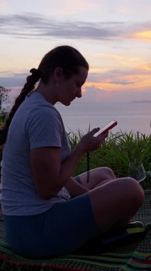 Dikey video. Mat 'te Oturan Kadın Okyanus, Geniş Açı, Rahat Yaşam Tarzı Konsepti Üzerinde Günbatımını izlerken Akıllı Telefon Kullanıyor