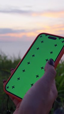 Dikey video. Deniz Altında Günbatımında Yukarı Kaydıran Yeşil Ekran ile Kadın El Tutma Akıllı Telefonu, Dijital Gevşeme Konsepti