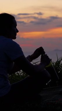 Dikey video. Mat 'in üzerinde oturan kadın, okyanus günbatımını izlerken şişeden şarap içiyor. Yan manzara, rahat yalnızlık kavramı.