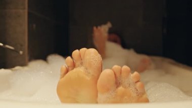 Ayaklar ön planda köpükle kaplıyken, kadın küvetin arkasında bulanıklaşırken alan etkisinin derinliğinde huzur ve rahatlığı vurgularken, Spa Dinlenme Konsepti