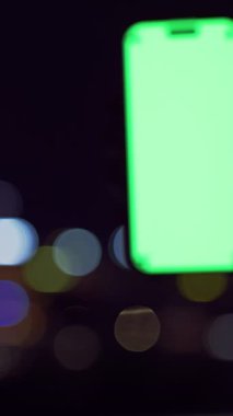 Dikey video. Yeşil Boş İzleme Ekranlı Akıllı Telefon Renkli Gece Şehri Bokeh 'e Karşı Yakın Açı Koyu Görünümlü Dijital Moda Anı Gösteriyor