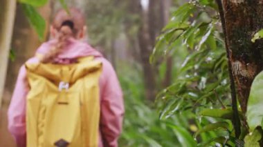 Pembe ceketli, sarı sırt çantalı bir kadın bulanık orman koridorunda yürüyor. Yumuşak odaklı bir resimde yemyeşil bitkiler ve soluk renkleri gösteriyor.