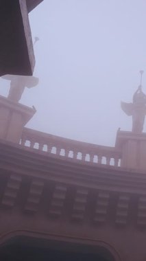 Dikey video. Ba Na Tepeleri 'nde düşük açılı, yumuşak soluk gökyüzünü ve sessiz mimari ayrıntıları gösteren yoğun sisin içinden yükselen melek heykelleri olan üst taştan bir bina.