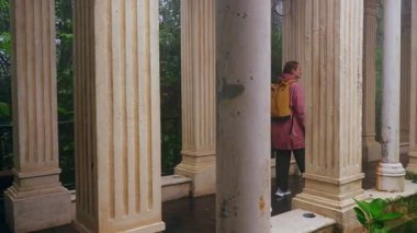 Sarı sırt çantalı kadın, Ba Na 'da uzun klasik sütunların arasında yürüyor. Geniş açılı çekimde, aşağıdaki sisli havayı ve yeşil bitkileri gösteriyor.