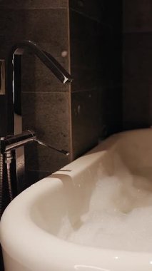 Vertical video. Foam Filled Bathtub in Dim Hotel Bathroom captured with Wide Angle and Warm Glow expressing Relaxation and Wellness Concept