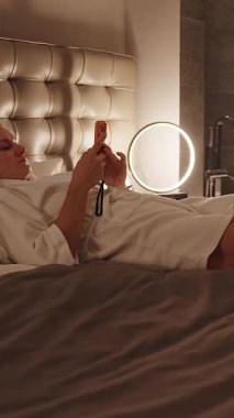 Dikey video. Bornozlu Otel Yatağında Rahatlayan Kadın Hafif Hafif ve Orta Yol Açısı ile Telefonu Kullanarak Konforlu Akşam Dinlenme Konsepti