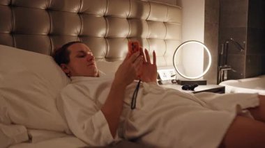 Woman Resting on Hotel Bed with Legs Extended and Phone in Hand captured with Low Angle and Soft Warm Light expressing Cozy Evening Comfort Concept
