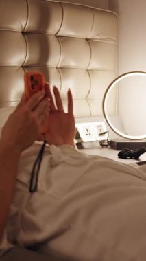 Dikey video. Otel Yatağında Dinlenen Bacakları Uzatılmış ve Telefonu Düşük Açı ve Yumuşak Işıltılı Gece Konforlu Konsepti ile Çekilen Kadın