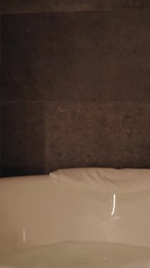 Dikey video. Otel Yatağında Dinlenen Kadın Işıklı Banyo ve Küvet 'in yanındaki telefonu kullanıyor. Düşük Açı ve Sıcak Ortam Işığı ile yakalanmış.