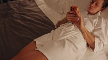 Bornozlu Kadın Bacakları Uzatılmış Otel Yatağında Dinleniyor Düşük Açı ve Yumuşak Ilık Işık ile çekilen Telefonu kullanarak Rahat Akşam Dinlenme Konsepti